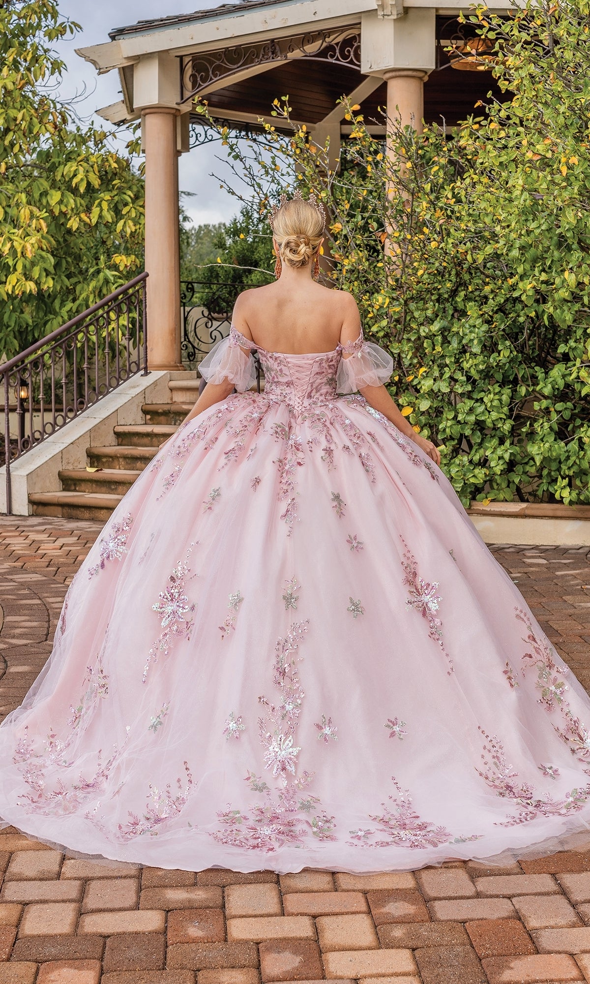 A woman wearing the Quinceanera Dress 1821 by Dancing Queen, a blush pink off-the-shoulder ball gown with floral embroidery, stands outdoors facing away before stone steps and lush greenery.